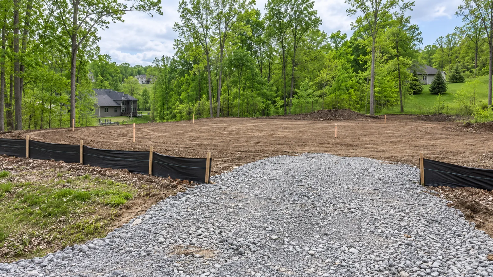 Cleared building lot with silt fence, a stone construction entrance, and corner stakes on a wooded lot in late spring
